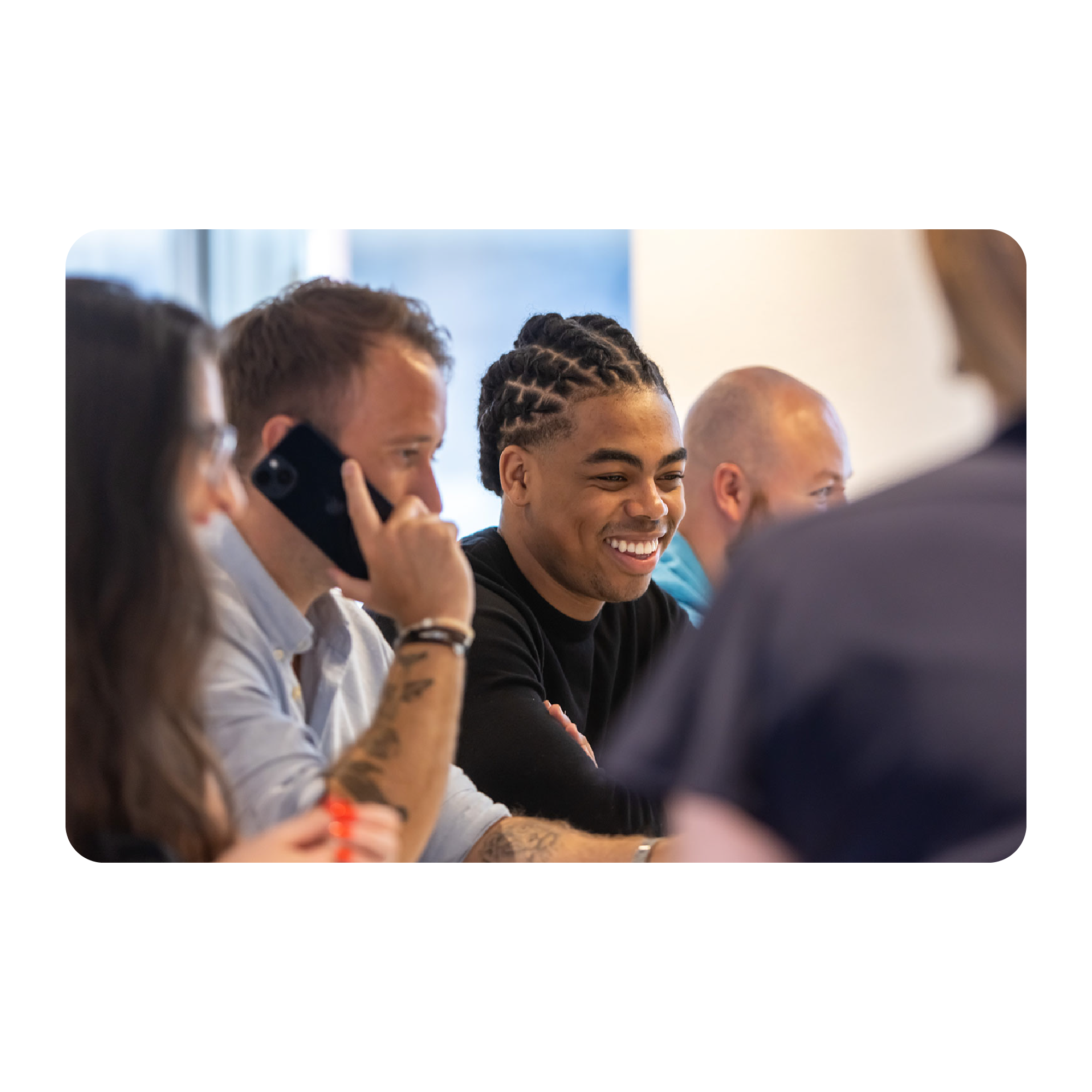 4 employees are pictured in frame with 2 in foreground out of focus. One male employee is holding a smart phone to his ear and the other is seated beside him, smiling