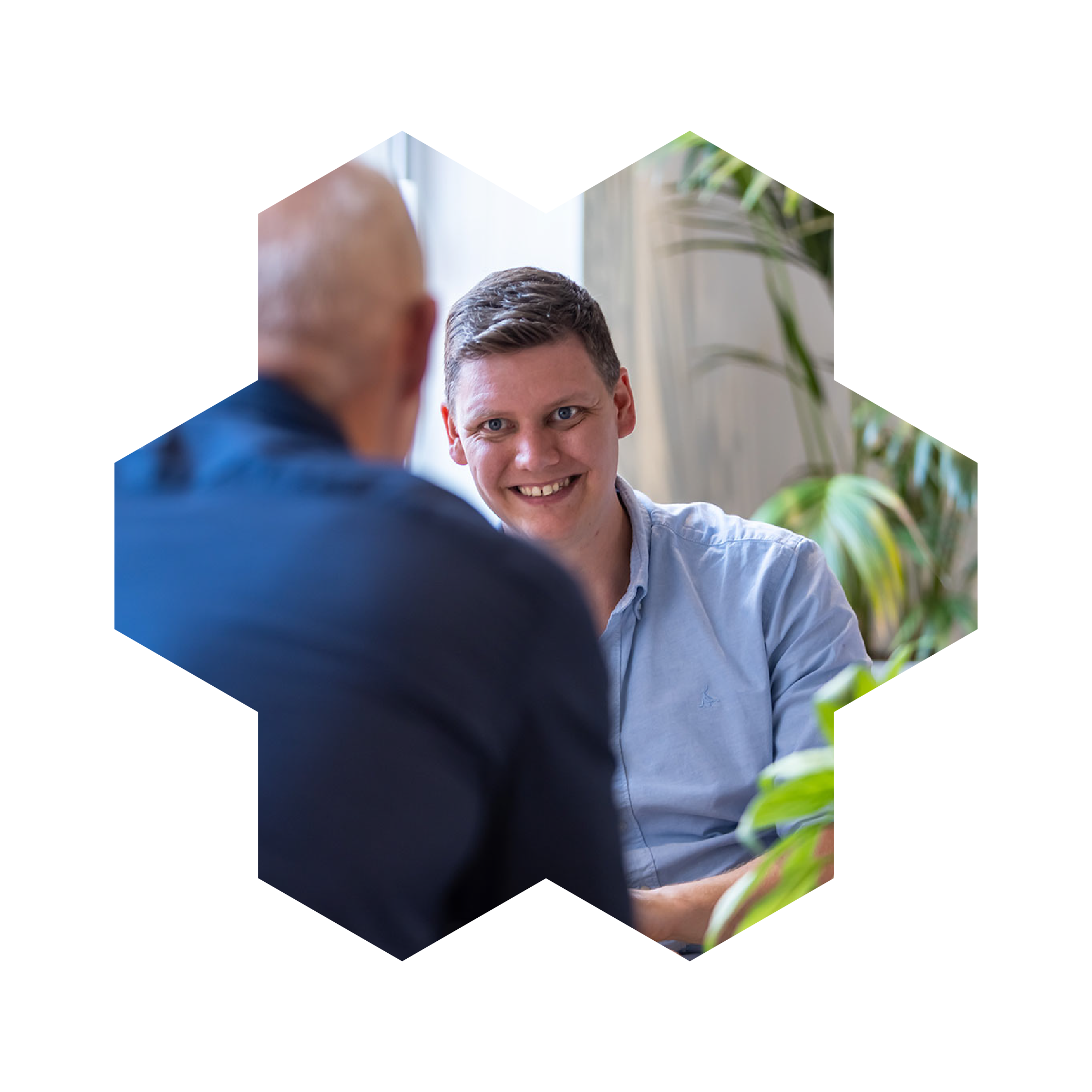 Two male colleagues in conversation. One colleague in the foreground has his back to the camera and is sitting directly opposite the colleague in the background.