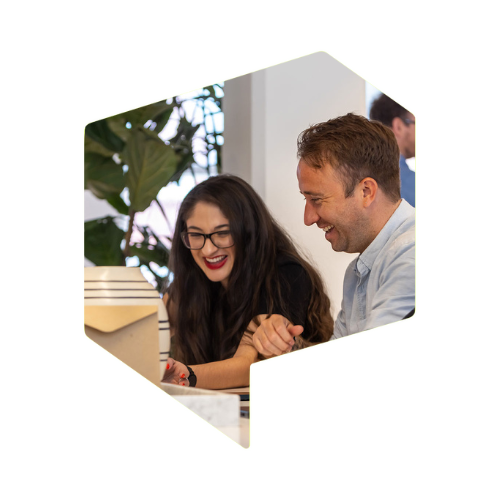 A woman and a man are sat at a meeting table smiling at a laptop