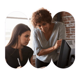 A woman sitting at a computer with her pen in hand is accompanied by her mentor, a woman who is stood next to her pointing at the monitor