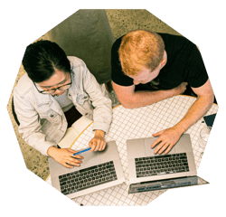 An aerial shot of a man and a woman sitting at a desk collaborating with their laptops