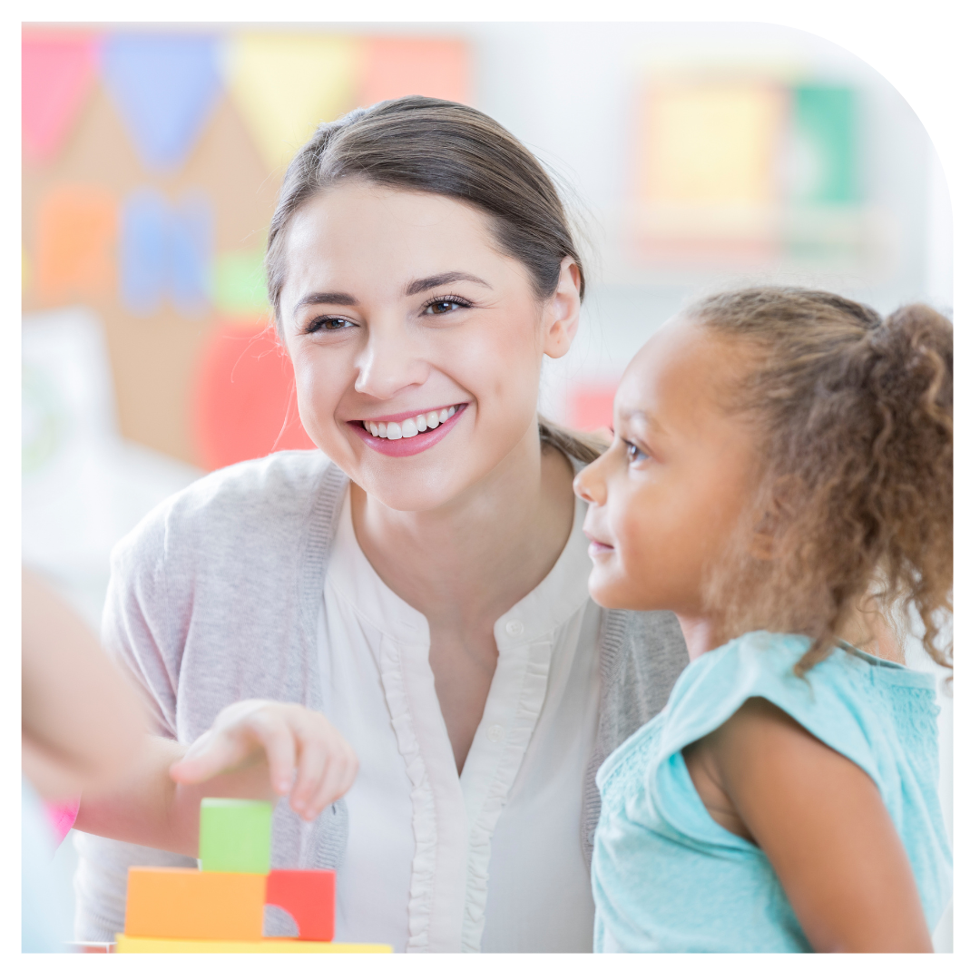 Preschool teacher is smiling at a child off camera and a girl in a blue tshirt is standing next to her