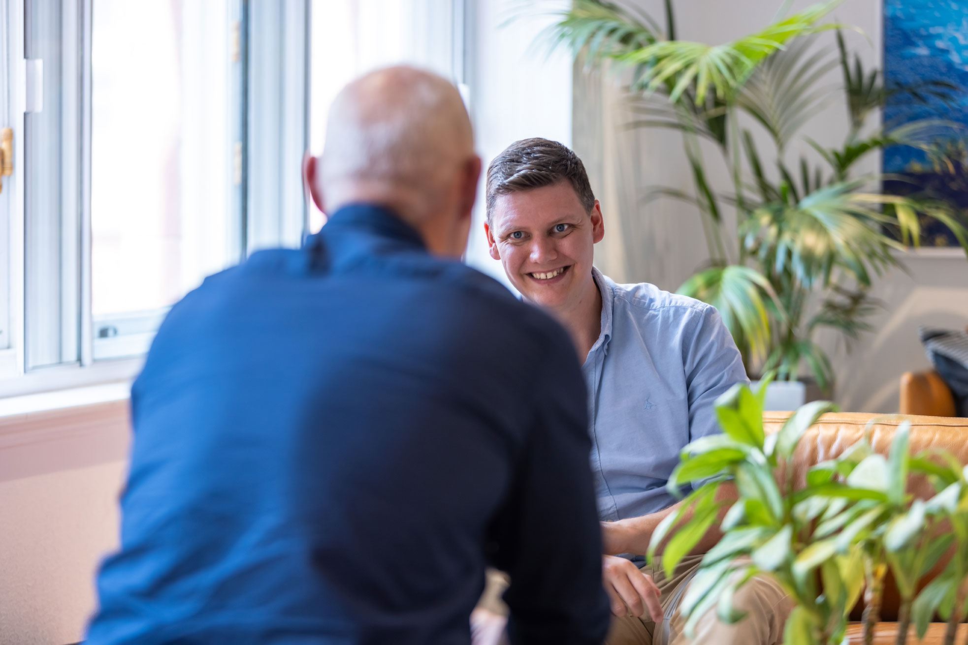 Two male colleagues in conversation. One has his back to the camera and is sitting directly opposite his colleague who is smiling.