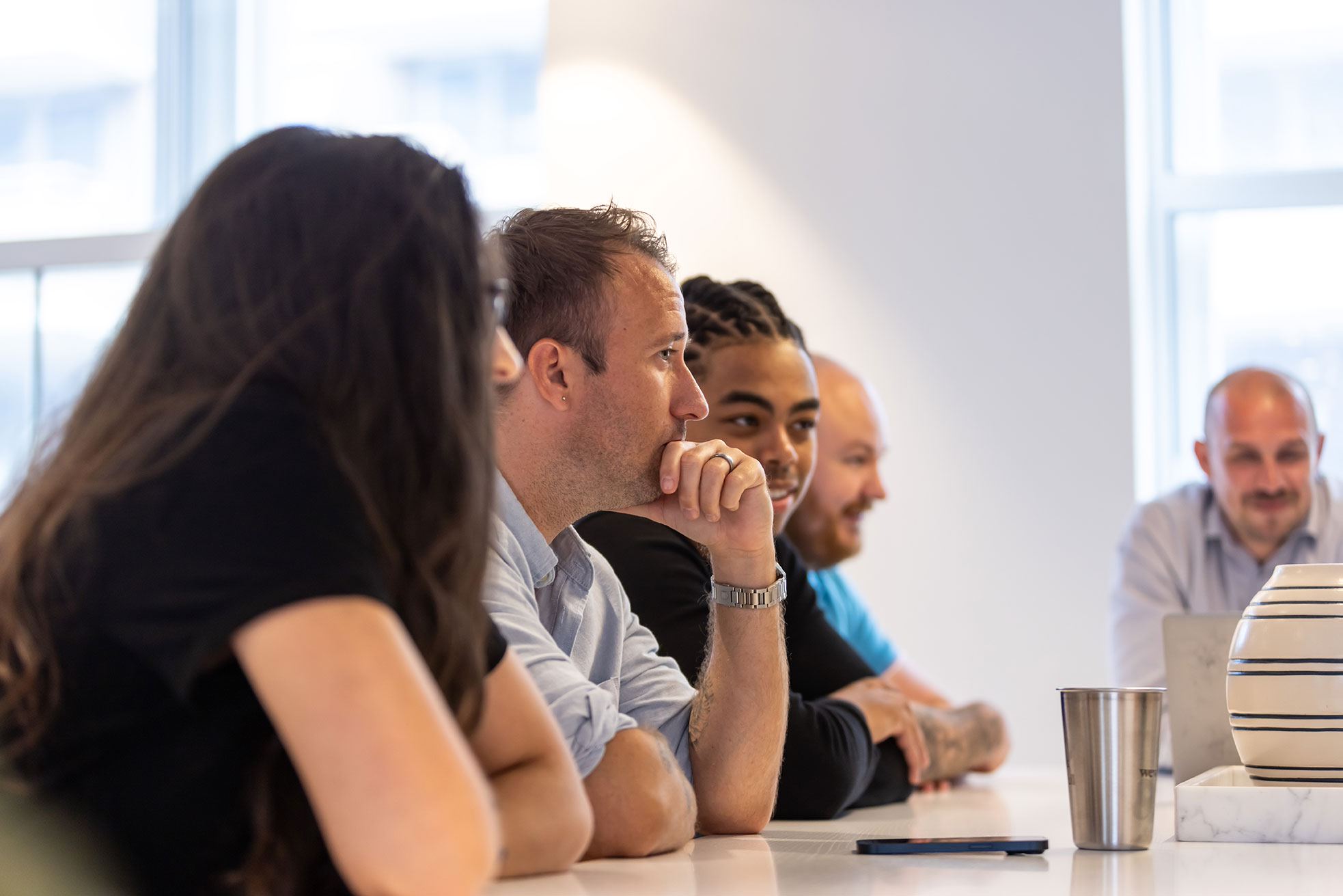 A man is sitting among 3 other colleagues who out of focus. They are in a meeting and he is listening intently.