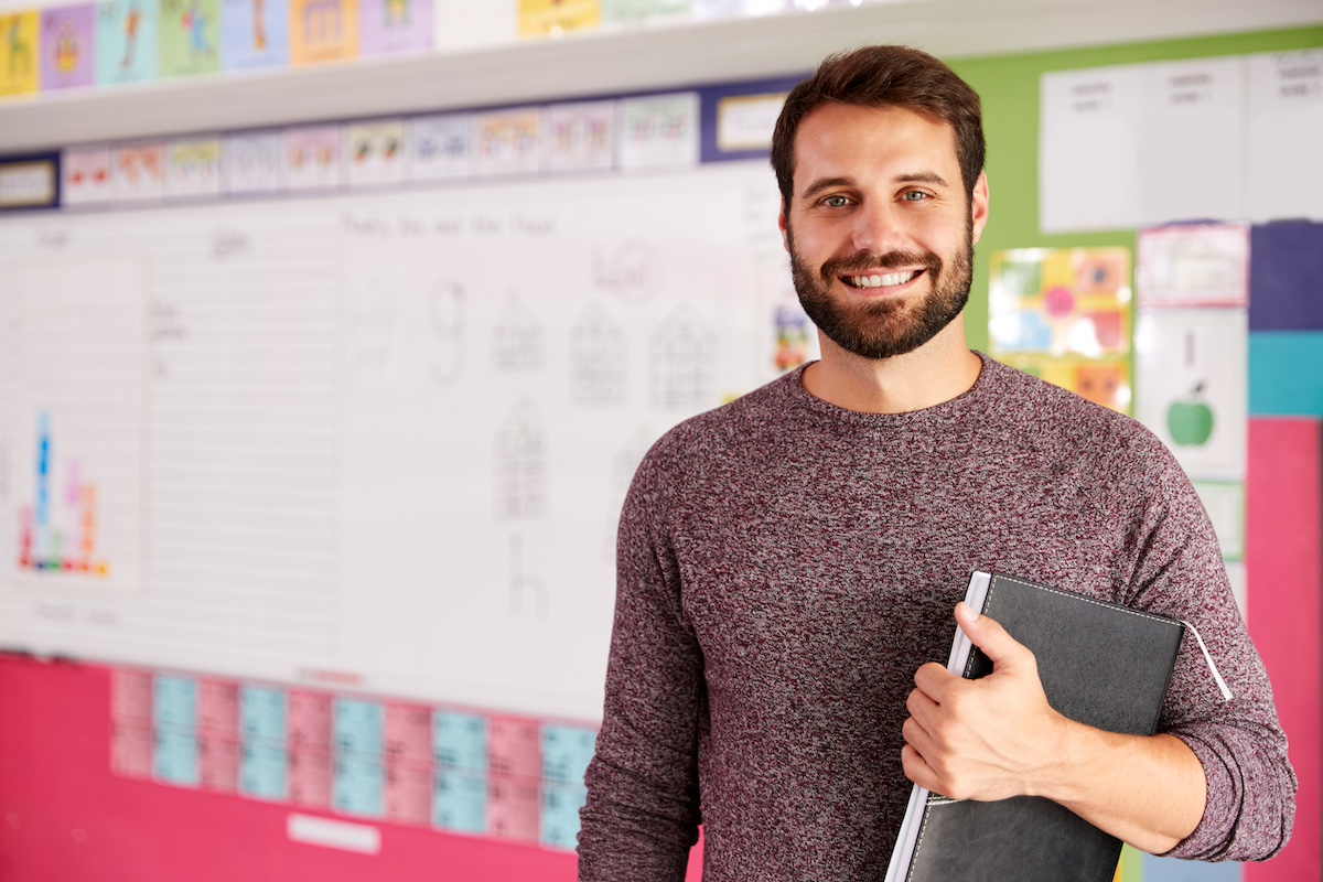 A male teacher is standing in front of a white board holding a folder and smiling