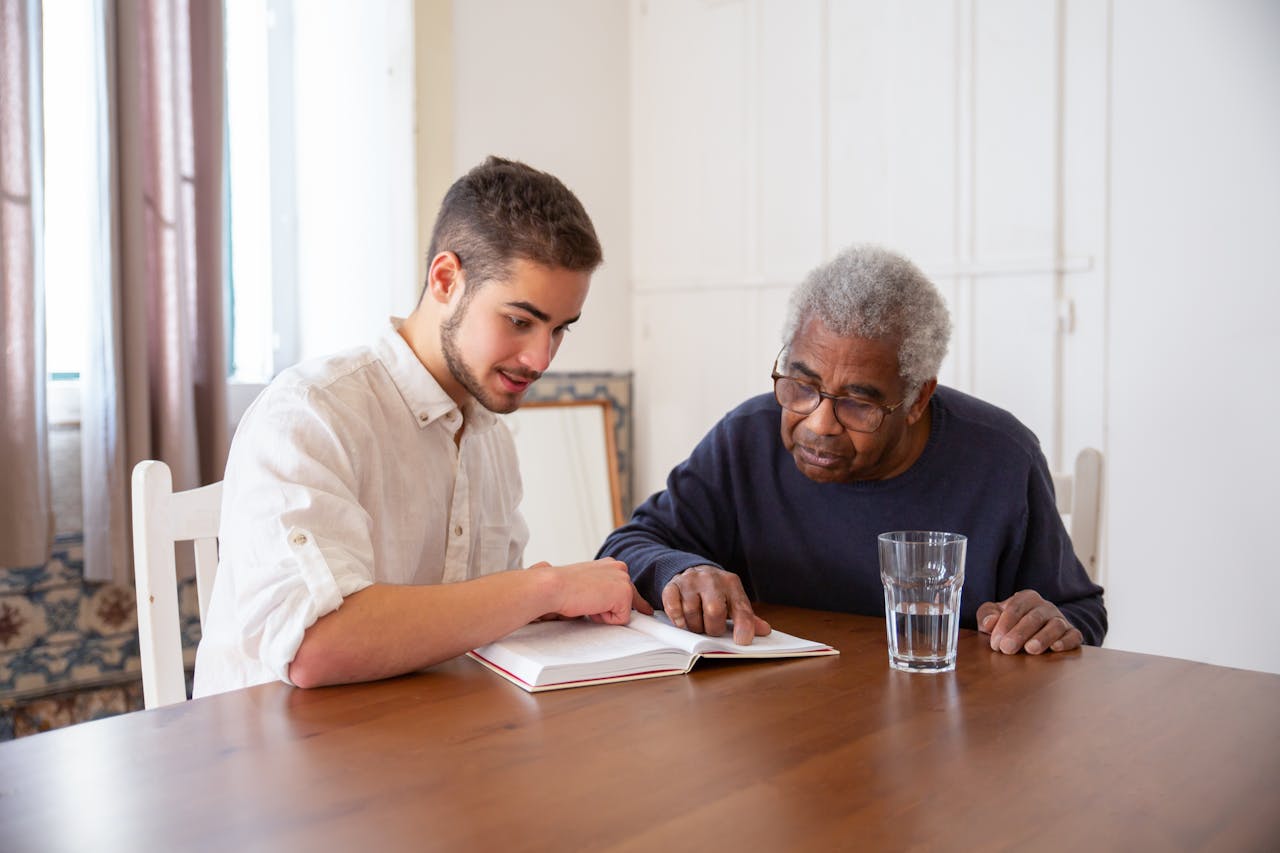 Male support worker reading a book at a dining table with an elderly male resident